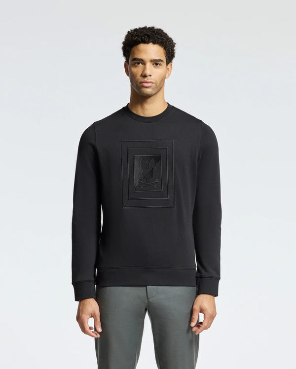 A person wearing the Psycho Bunny MENS OAK CITY CREW NECK - B6S274E200 sweatshirt, featuring raised embroidery and paired with gray pants, stands against a plain light background. The lightweight crew neck design and premium quality fabric enhance the subtle yet stylish appearance.