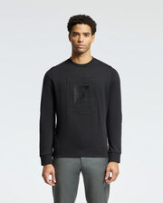 A person wearing the Psycho Bunny MENS OAK CITY CREW NECK - B6S274E200 sweatshirt, featuring raised embroidery and paired with gray pants, stands against a plain light background. The lightweight crew neck design and premium quality fabric enhance the subtle yet stylish appearance.