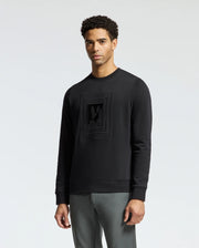 A man wearing the Psycho Bunny MENS OAK CITY CREW NECK - B6S274E200, which is a lightweight sweatshirt with premium quality raised embroidery on the front, stands against a plain light background. He has short curly hair and is dressed in gray pants.