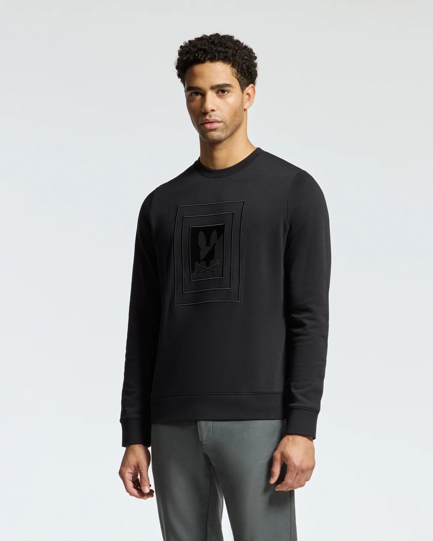A man wearing the Psycho Bunny MENS OAK CITY CREW NECK - B6S274E200, which is a lightweight sweatshirt with premium quality raised embroidery on the front, stands against a plain light background. He has short curly hair and is dressed in gray pants.