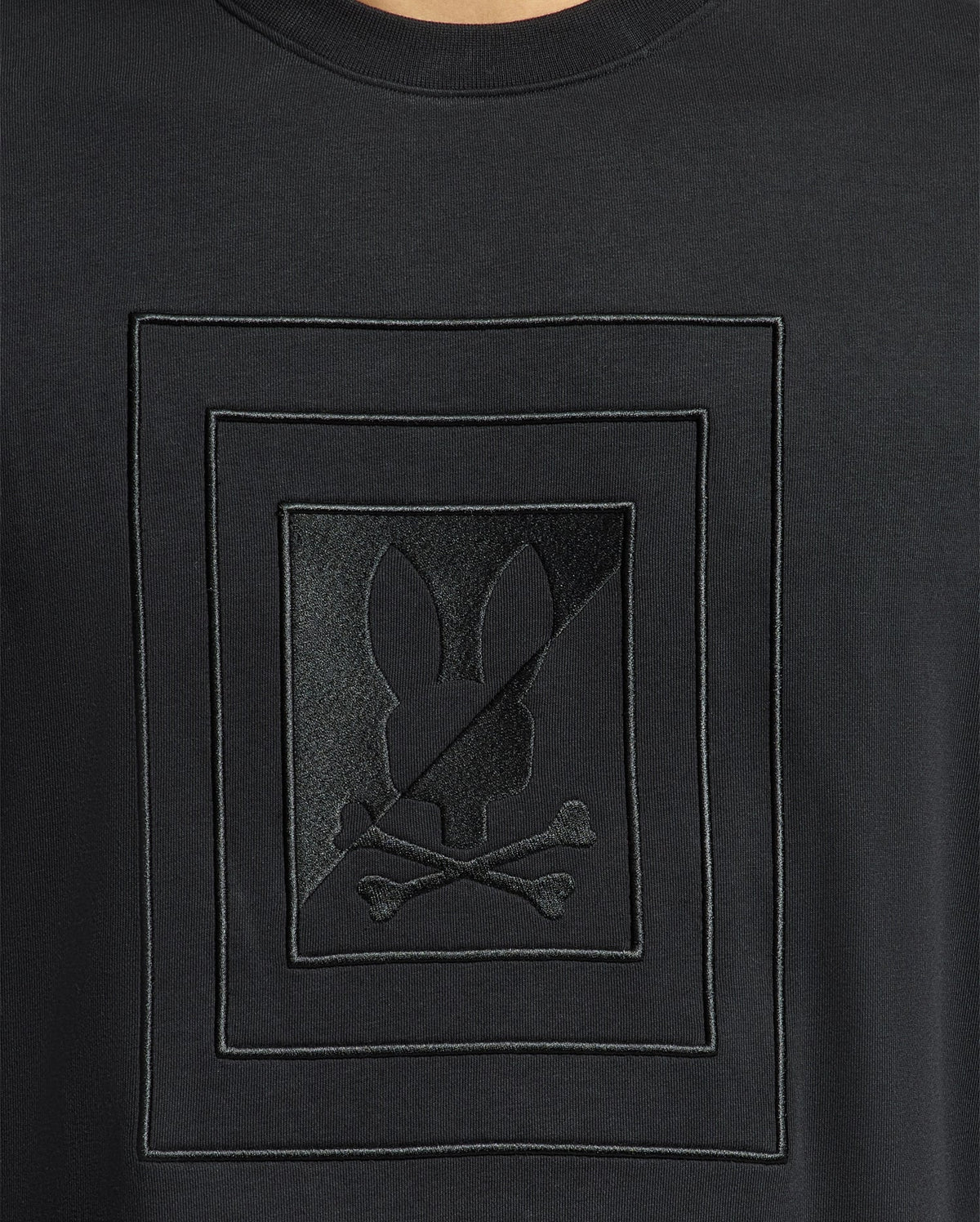 Close-up of the Psycho Bunny black shirt, MENS OAK CITY CREW NECK - B6S274E200, crafted from premium quality fabric and featuring a distinctive embossed design: a silhouette of a rabbit head above crossed bones, framed within geometric borders. This lightweight crew neck shirt is enhanced with raised embroidery for added texture and style.