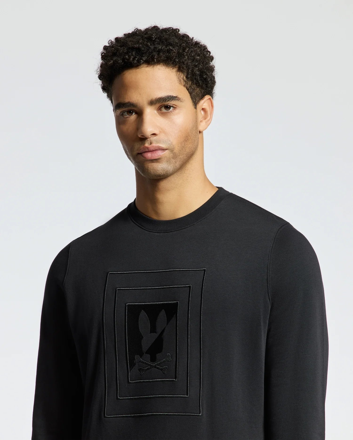A person with curly hair is wearing the Psycho Bunny MENS OAK CITY CREW NECK - B6S274E200, a lightweight black sweater featuring a rectangular pattern with abstract designs. The premium quality fabric stands out against the plain, light grey background.