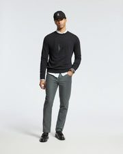 A man stands against a plain background, wearing a black cap and the MENS OAK CITY CREW NECK from Psycho Bunny over a white shirt, paired with gray pants and black loafers. He has one hand in his pocket and wears a watch on his wrist.