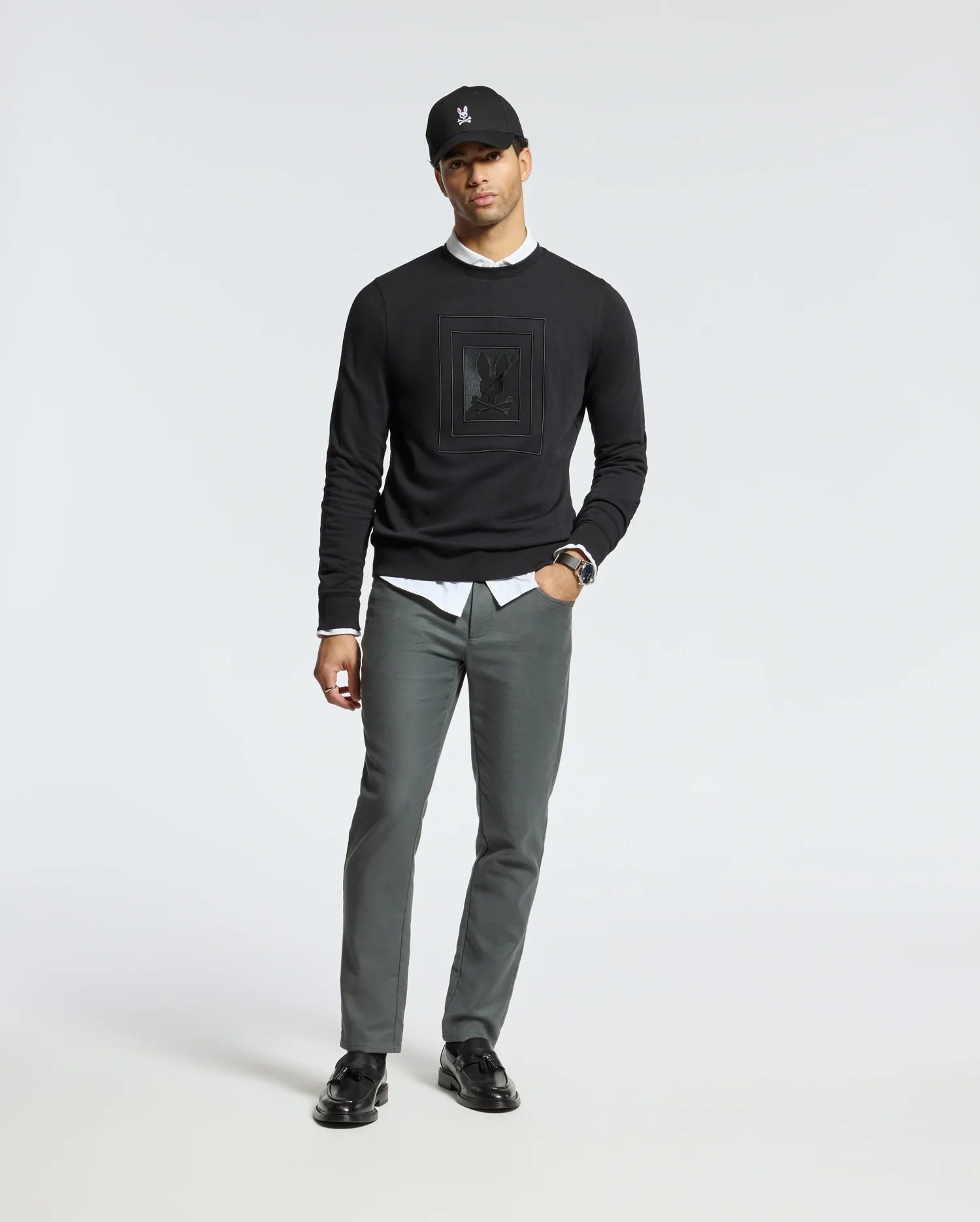 A man stands against a plain background, wearing a black cap and the MENS OAK CITY CREW NECK from Psycho Bunny over a white shirt, paired with gray pants and black loafers. He has one hand in his pocket and wears a watch on his wrist.