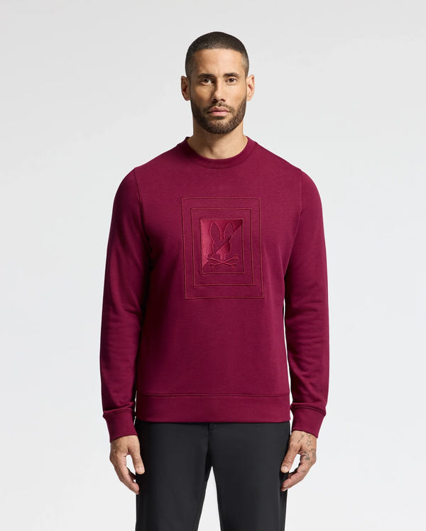 A man with short hair and a beard stands against a plain white background, showcasing the Psycho Bunny MENS OAK CITY CREW NECK - B6S274E200 in burgundy. This premium quality piece features an embossed design on the front, perfectly paired with black pants.