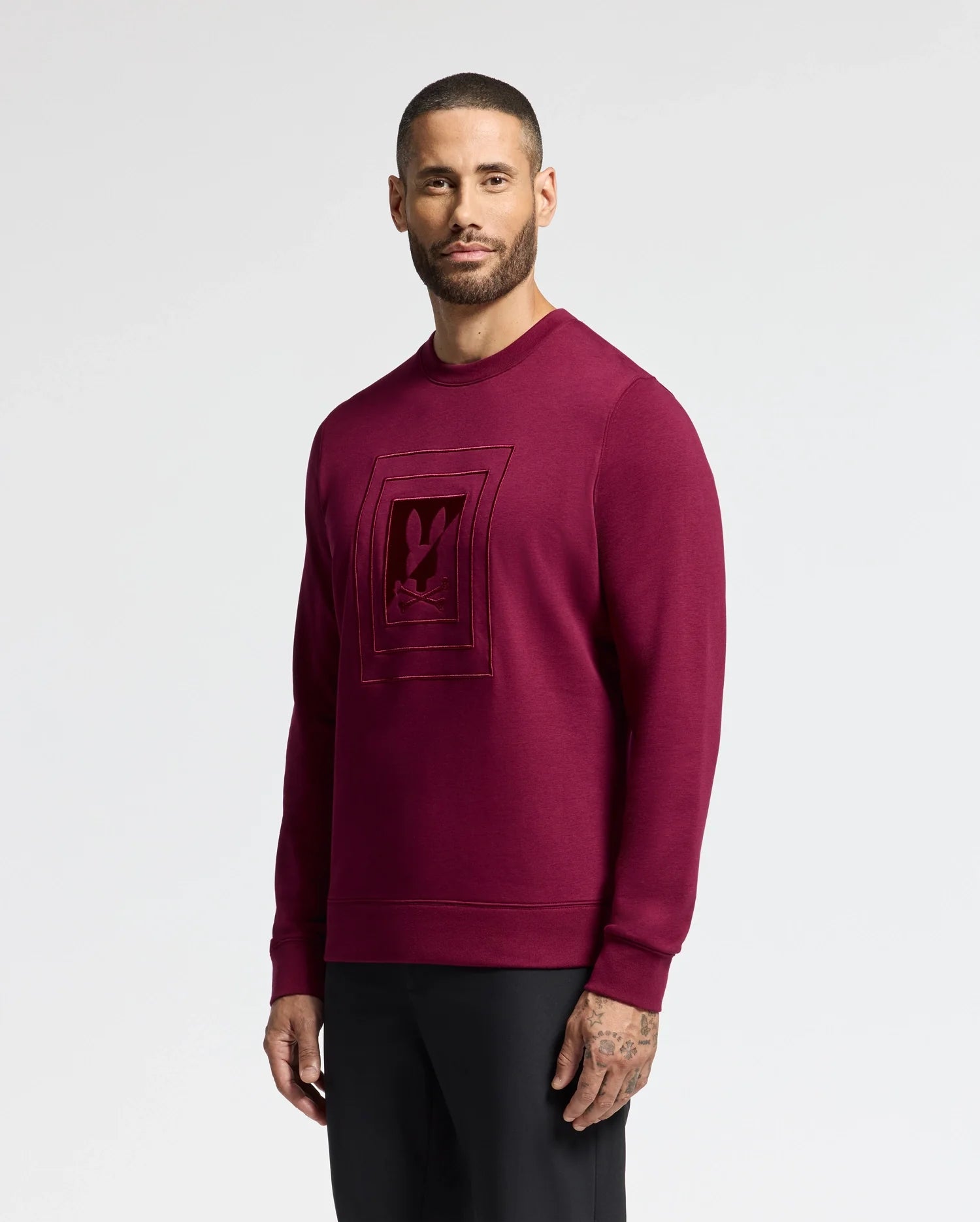 A man in a Psycho Bunny MENS OAK CITY CREW NECK - B6S274E200 stands against a plain white background. Sporting short hair and a beard, he looks directly at the camera while pairing his premium quality top with black pants.