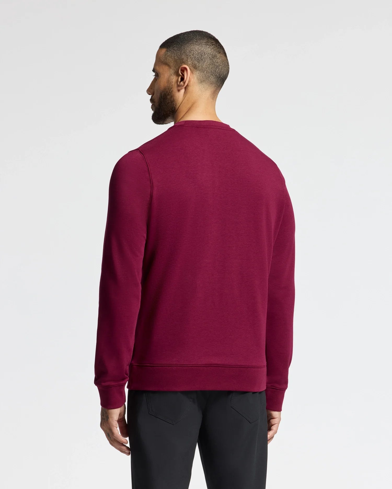 A man with short dark hair and a beard is seen from the back, wearing the Psycho Bunny MENS OAK CITY CREW NECK - B6S274E200 sweater in a dark red hue and paired with black pants. The plain white background accentuates the elegance of the sweater's raised embroidery.