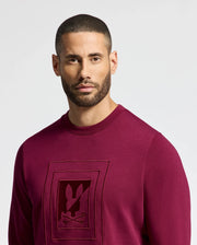 A man with a buzz cut and beard is dressed in a Psycho Bunny MENS OAK CITY CREW NECK - B6S274E200 sweater, which features premium quality maroon fabric and raised embroidery on the front. The background is plain white.