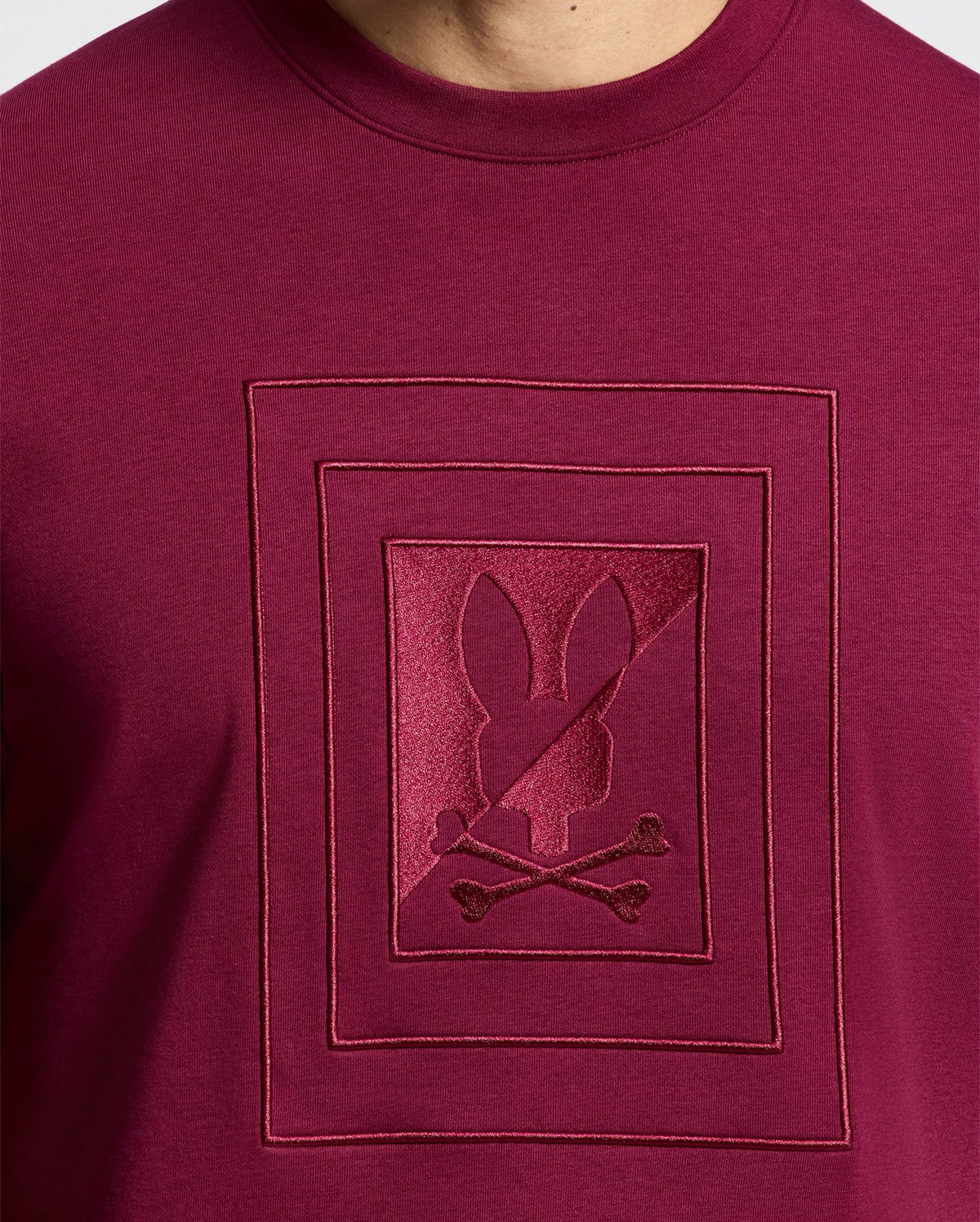 A close-up of the MENS OAK CITY CREW NECK - B6S274E200 by Psycho Bunny showcases its premium quality, featuring a raised embroidery design of a bunny silhouette above crossed bones, enclosed within two rectangular borders. The intricate stitching adds texture to the smooth maroon fabric.
