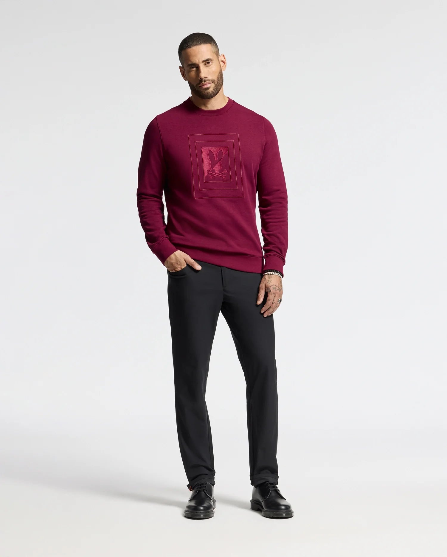 A person is positioned against a simple backdrop, dressed in the MENS OAK CITY CREW NECK - B6S274E200 sweater from Psycho Bunny. The sweater is dark red and features raised embroidery. They are paired with black pants and black shoes. The individual has a short haircut and a beard, with their left hand casually placed in their pocket.