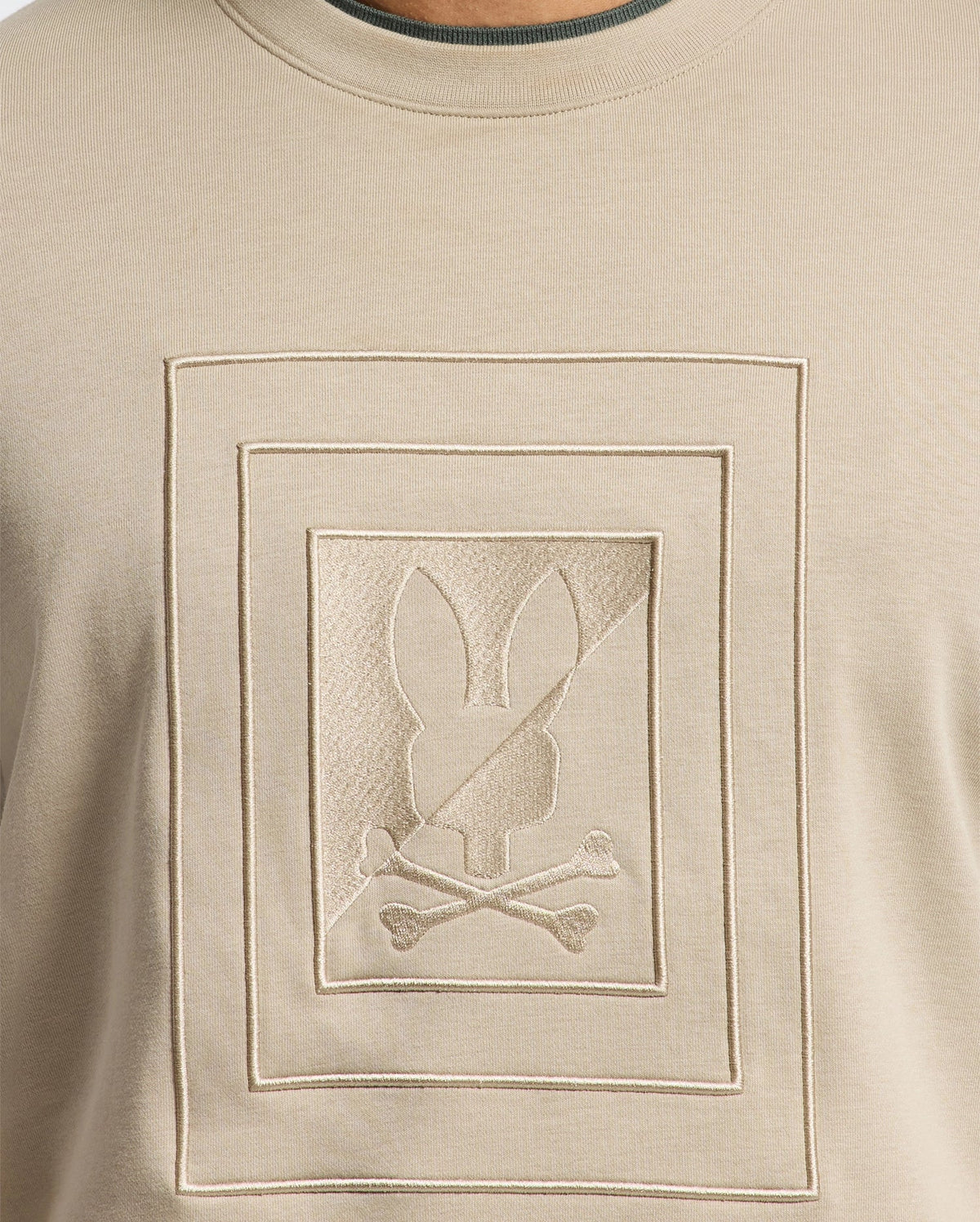 The Psycho Bunny MENS OAK CITY CREW NECK - B6S274E200 is a premium quality beige men's sweater that showcases a raised embroidery of an embossed rectangular design. Within the rectangle, there is a bunny head with crossbones beneath it. The subtle texture highlights contribute to its monochromatic, minimalist appearance.