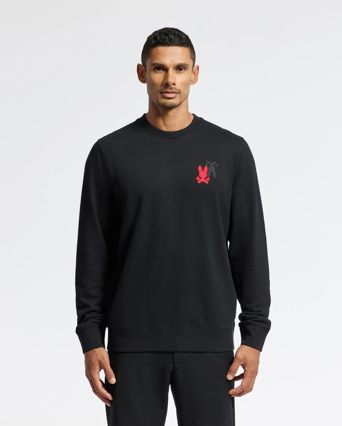 A person wearing the Psycho Bunny MENS PIERCE PEACHED TERRY CREW - B6S278E200 stands against a plain white background. The black sweatshirt is made from high-quality cotton and features intricate embroidery, long sleeves, a crew neck, and a small red logo on the chest. The individual has short, dark hair and an expressionless face.