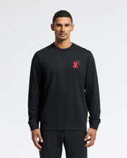 A person wearing the Psycho Bunny MENS PIERCE PEACHED TERRY CREW - B6S278E200 stands against a plain white background. The black sweatshirt is made from high-quality cotton and features intricate embroidery, long sleeves, a crew neck, and a small red logo on the chest. The individual has short, dark hair and an expressionless face.