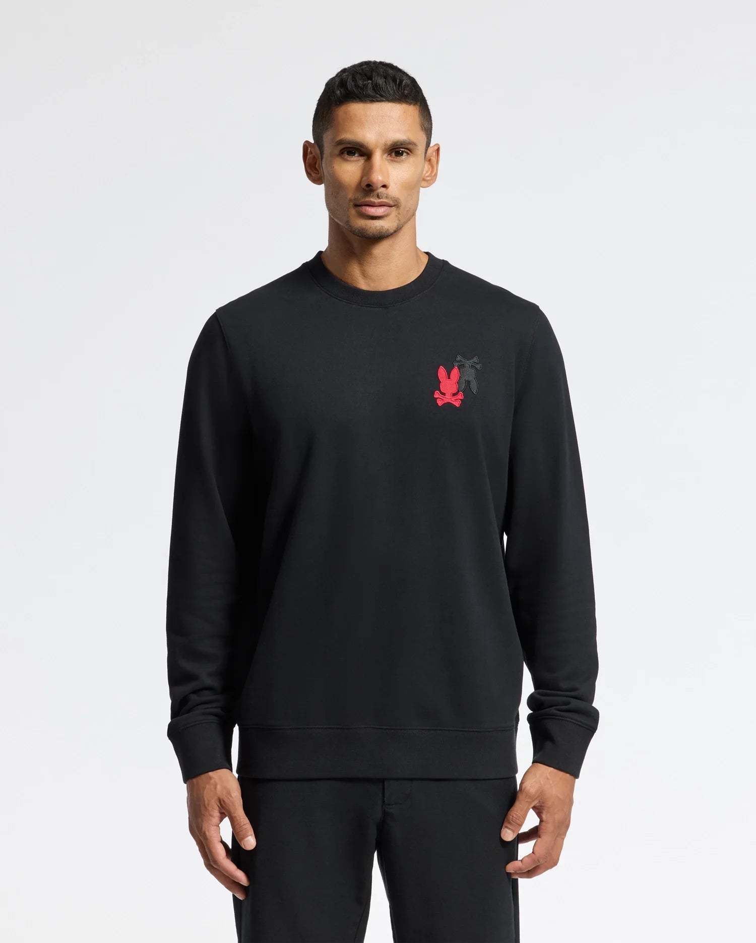 A person wearing the Psycho Bunny MENS PIERCE PEACHED TERRY CREW - B6S278E200 stands against a plain white background. The black sweatshirt is made from high-quality cotton and features intricate embroidery, long sleeves, a crew neck, and a small red logo on the chest. The individual has short, dark hair and an expressionless face.