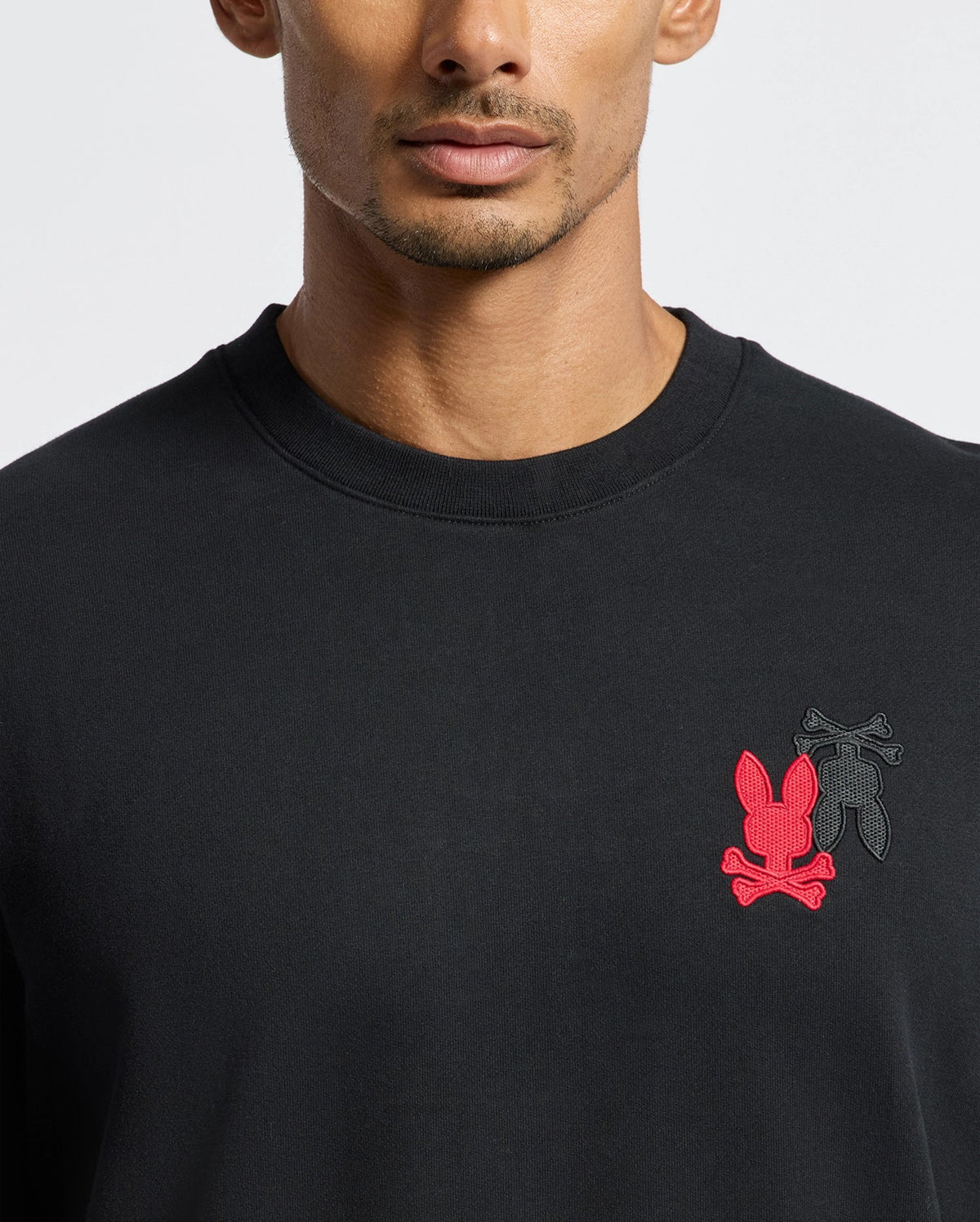 The image showcases a man wearing the stylish MENS PIERCE PEACHED TERRY CREW - B6S278E200 from Psycho Bunny. This sophisticated black crew-neck sweatshirt, crafted from premium cotton, is adorned with two embroidered logos: a red bunny head with crossbones and an overlaying black one. The photo frames him from his shoulders to his head as he faces forward.
