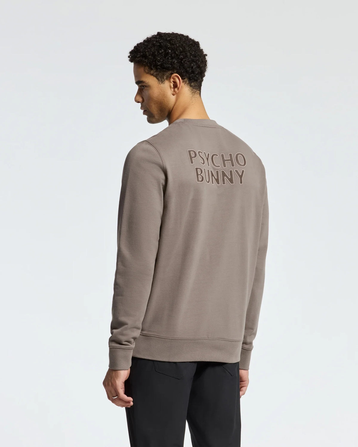 A person with short curly hair is shown from behind, wearing the MENS PIERCE PEACHED TERRY CREW - B6S278E200 by Psycho Bunny. This stylish beige sweatshirt pairs effortlessly with their black pants and features a plain, light-colored background.