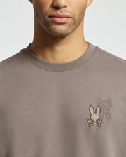 A man wearing the Psycho Bunny MENS PIERCE PEACHED TERRY CREW sweater in taupe, featuring an embroidered logo with bunny silhouettes and crossed bones on the chest, made of high-quality cotton.