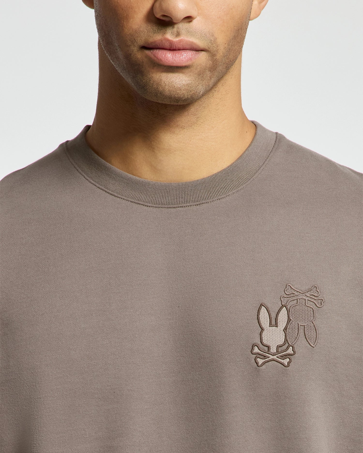 A man wearing the Psycho Bunny MENS PIERCE PEACHED TERRY CREW sweater in taupe, featuring an embroidered logo with bunny silhouettes and crossed bones on the chest, made of high-quality cotton.