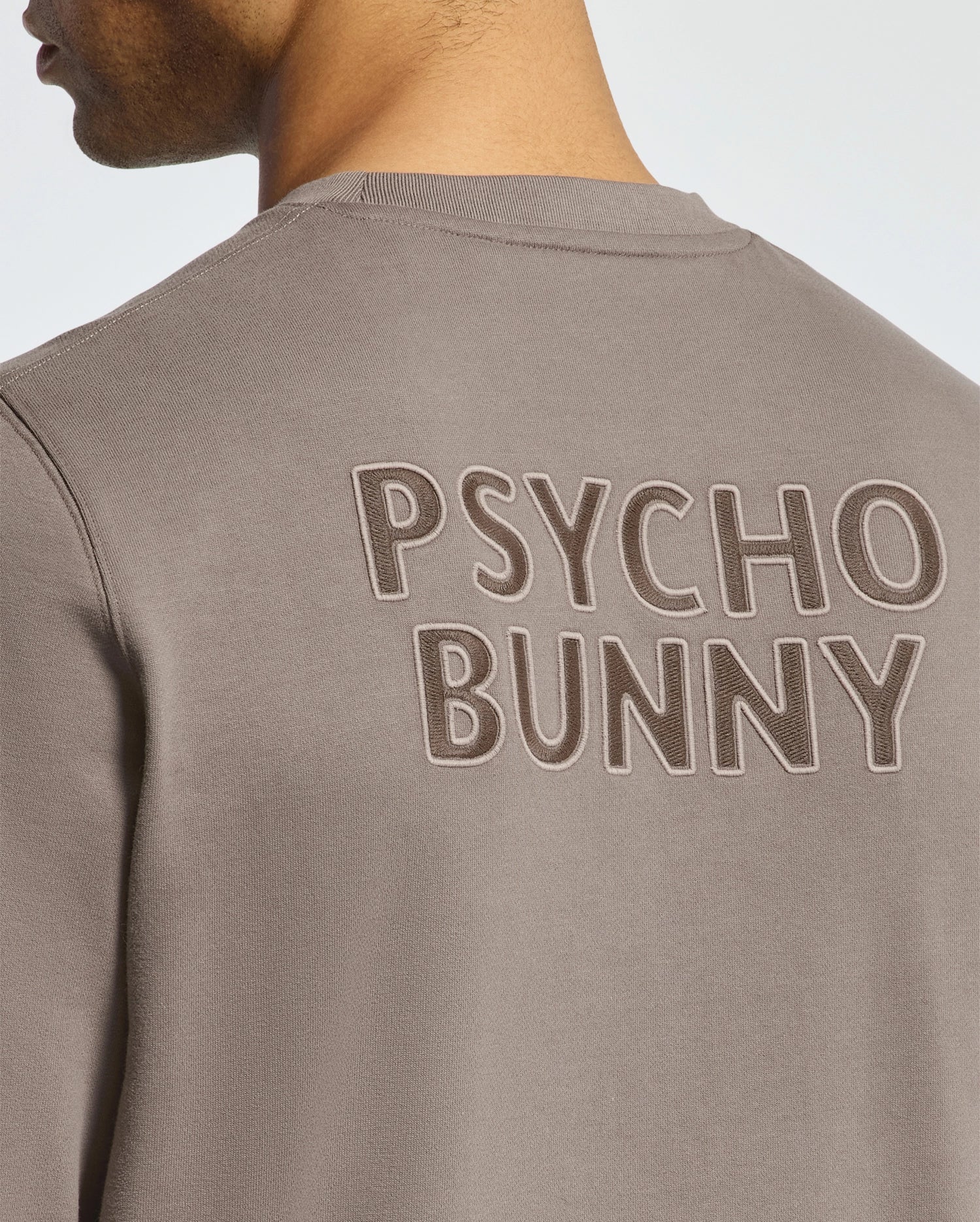 A person wears the Men's Pierce Peached Terry Crew by Psycho Bunny, featuring "PSYCHO BUNNY" in large capital letters on the back. Made from high-quality cotton, the focus is on the bold text, with only part of their head and shoulder visible.