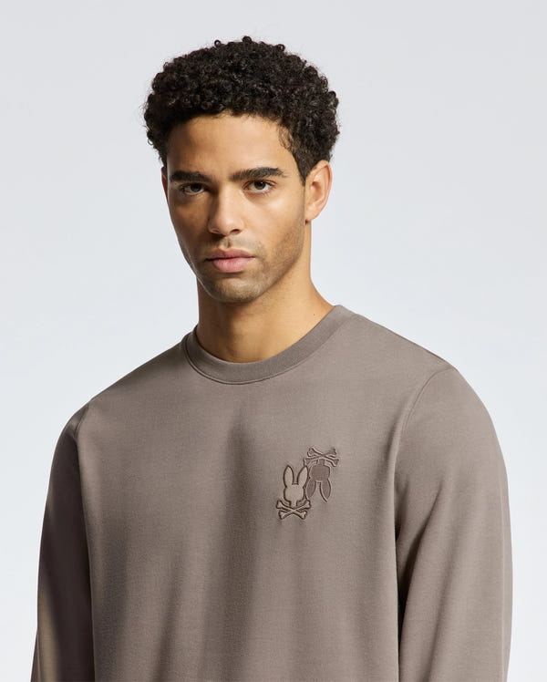 A man with short curly hair wears a Psycho Bunny MENS PIERCE PEACHED TERRY CREW (B6S278E200), made from premium cotton. The stylish light brown sweatshirt features an understated chest design as he poses against a plain gray background.