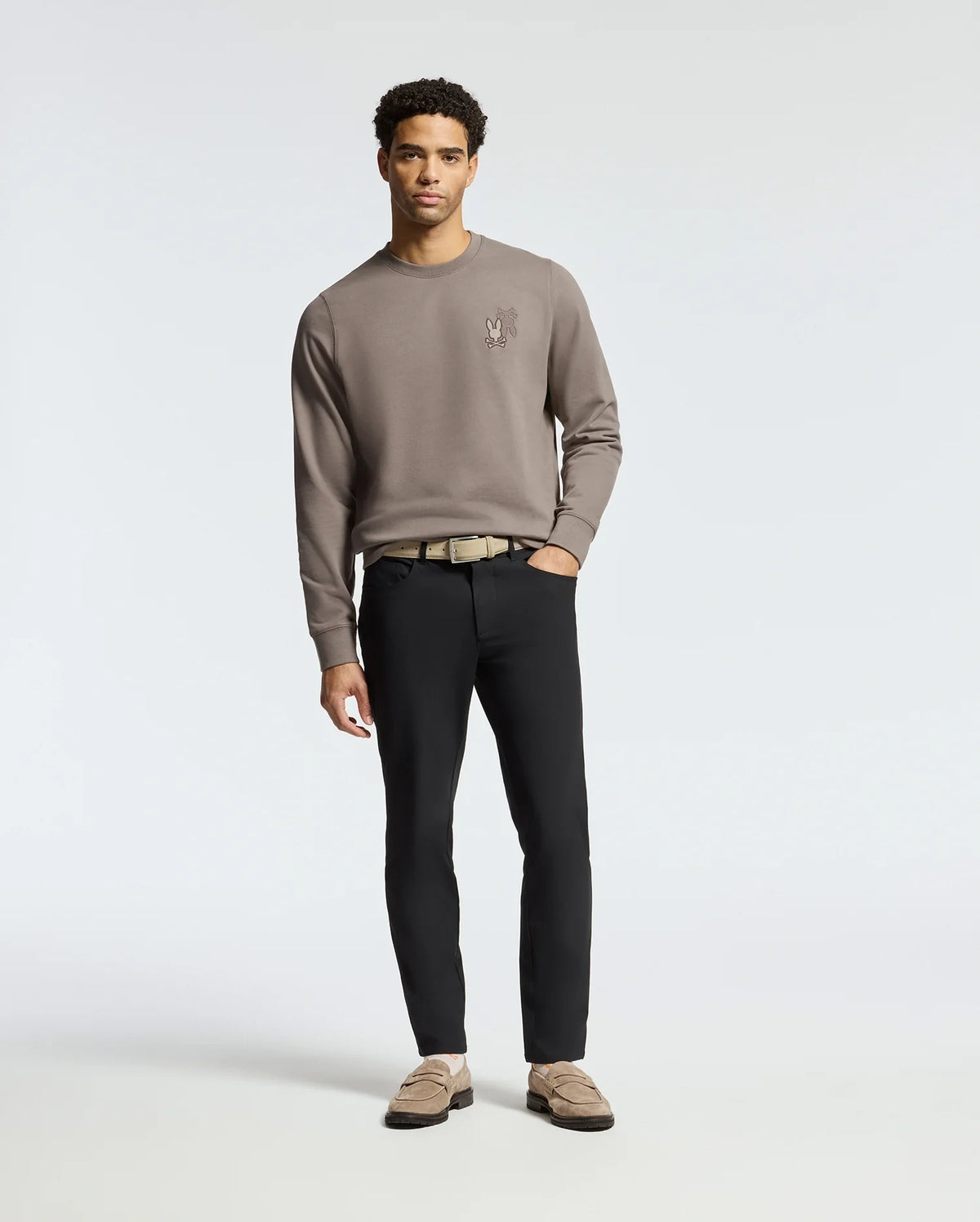 A man stands against a plain backdrop, wearing the Psycho Bunny MENS PIERCE PEACHED TERRY CREW sweatshirt in taupe with a chest design, paired with black pants and tan loafers. His hands at his sides, he gazes directly at the camera for an effortlessly stylish look.