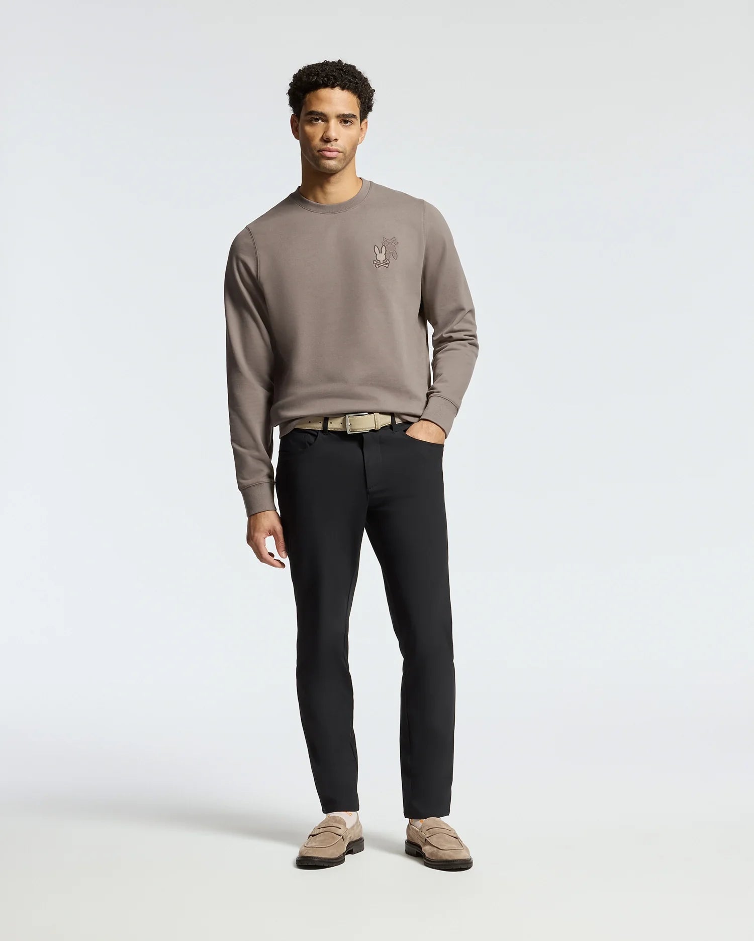 A man stands against a plain backdrop, wearing the Psycho Bunny MENS PIERCE PEACHED TERRY CREW sweatshirt in taupe with a chest design, paired with black pants and tan loafers. His hands at his sides, he gazes directly at the camera for an effortlessly stylish look.