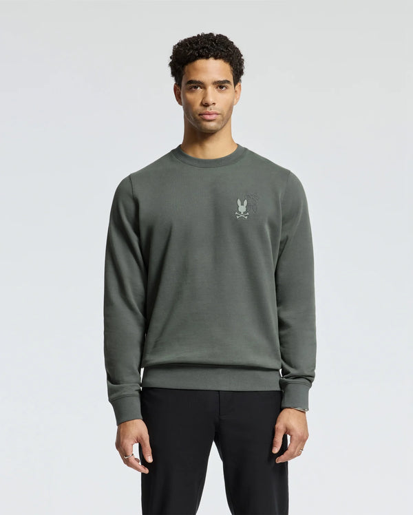 A person wearing the Psycho Bunny MENS WORDMARK PEACHED TERRY CREW in dark green, showcasing an embroidered Bunnies logo on the left chest, along with black pants, stands against a light gray background.