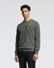A person wearing the Psycho Bunny MENS WORDMARK PEACHED TERRY CREW in dark green, which features an embroidered rooster emblem on the left chest, stands against a plain background. The knit cotton fabric harmonizes with their short, curly hair as they gaze directly at the camera.