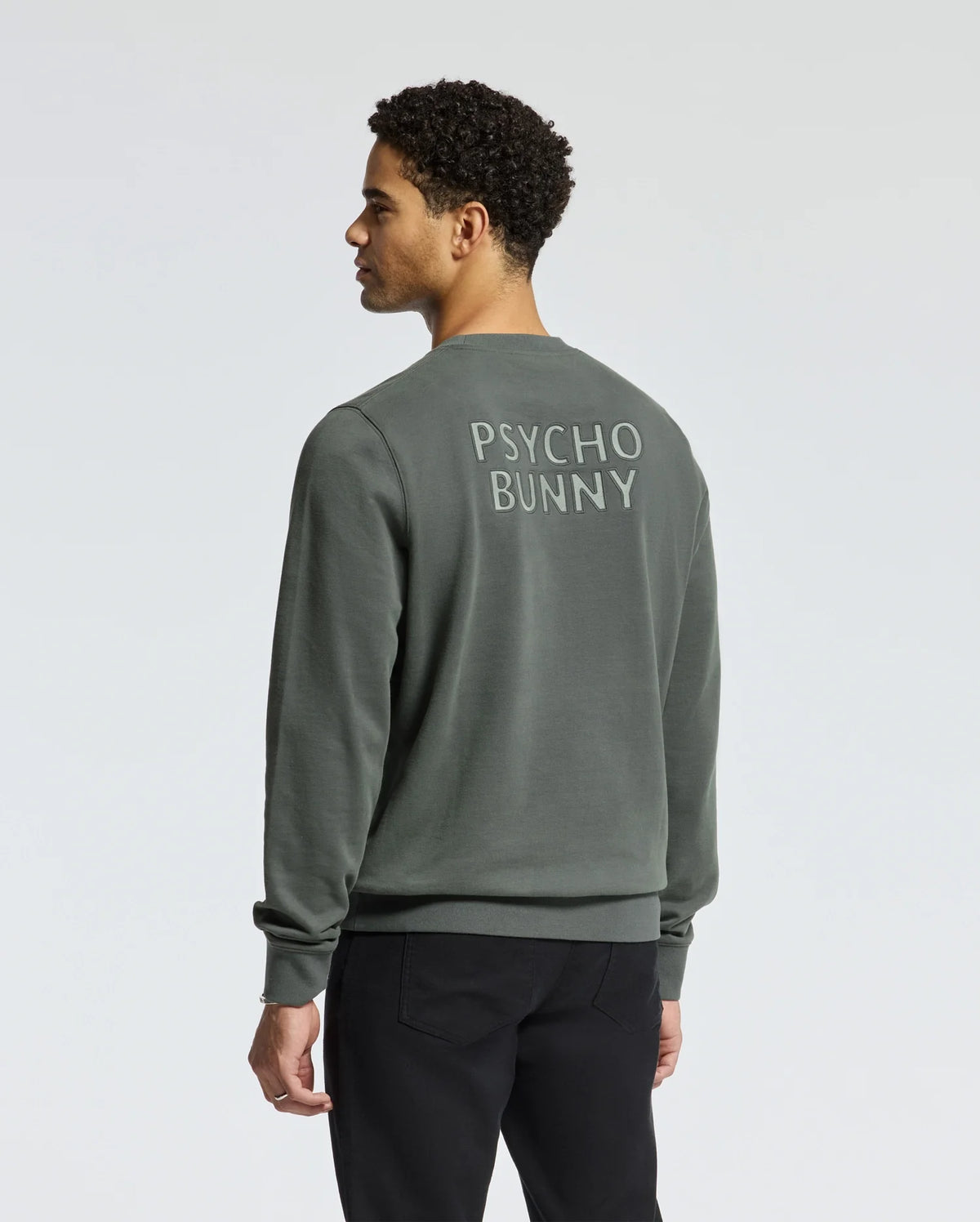 A man wearing the MENS WORDMARK PEACHED TERRY CREW - B6S278E200 by Psycho Bunny, which features a dark green knit cotton design with "Psycho Bunny" text on the back, stands against a plain background, facing away from the camera.