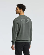 A man wearing the MENS WORDMARK PEACHED TERRY CREW - B6S278E200 by Psycho Bunny, which features a dark green knit cotton design with "Psycho Bunny" text on the back, stands against a plain background, facing away from the camera.