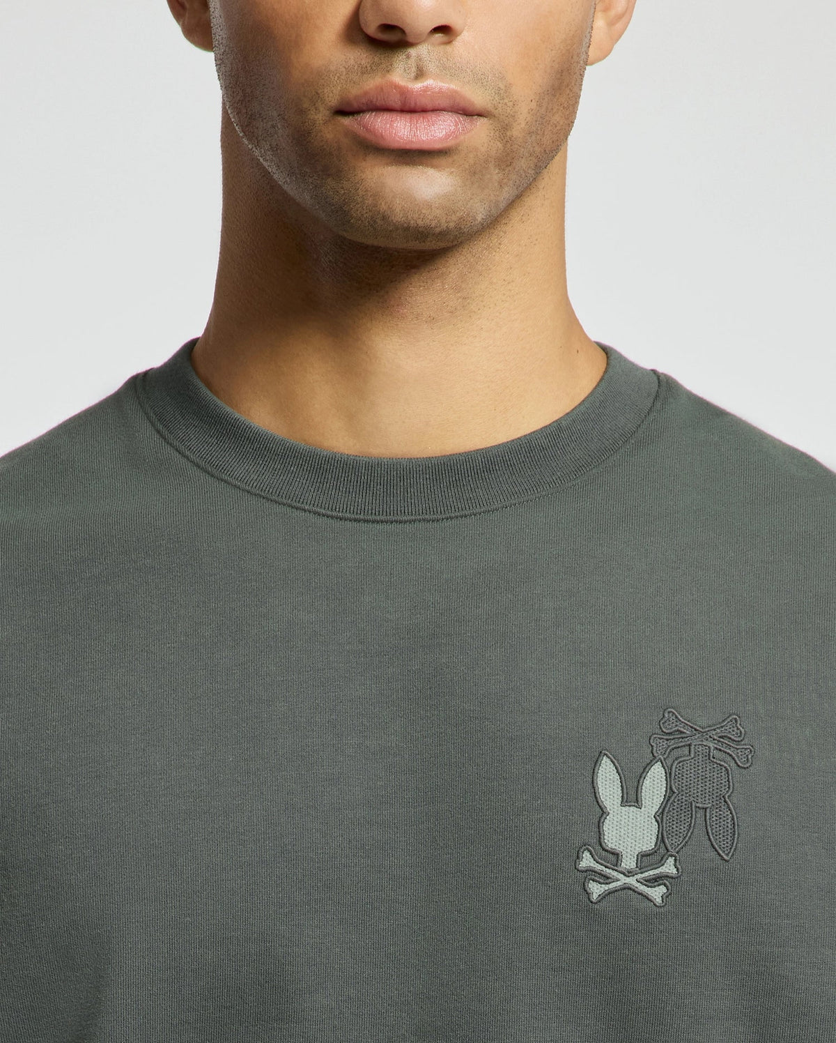 A man wears the Psycho Bunny MENS WORDMARK PEACHED TERRY CREW - B6S278E200, a dark green knit cotton pullover with an embroidered bunny and crossbones design on the chest set against a plain background.