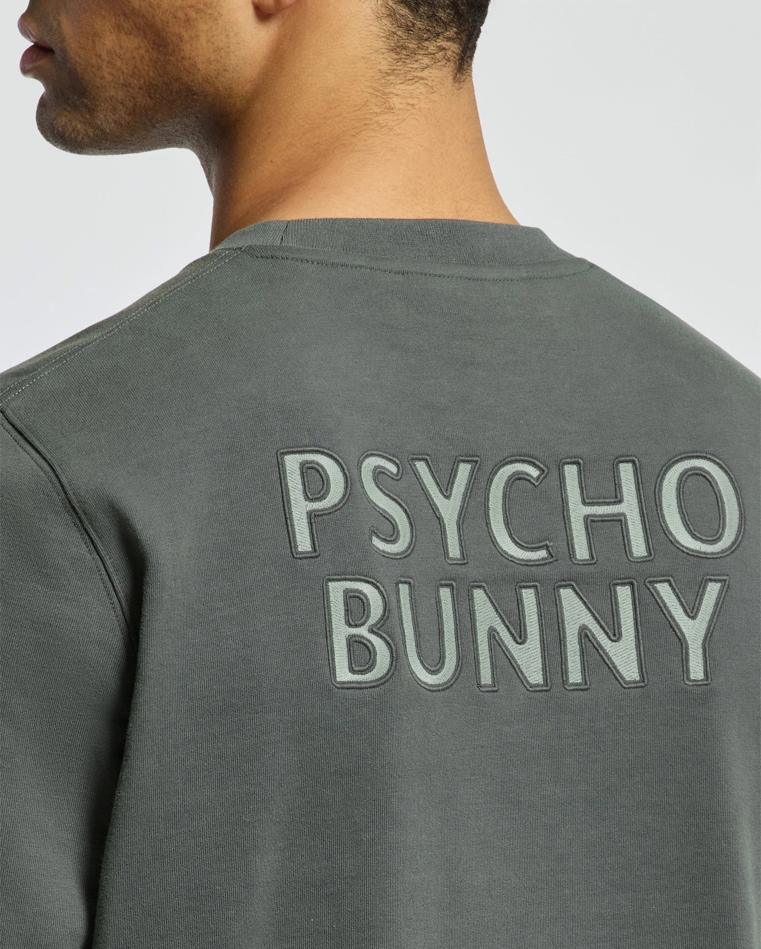 A person wearing the MENS WORDMARK PEACHED TERRY CREW - B6S278E200 from Psycho Bunny, showcasing a dark green knit cotton shirt with "PSYCHO BUNNY" prominently displayed across the back. The image features a side view emphasizing the upper back and neck area, highlighting the pullover's casual yet stylish design.