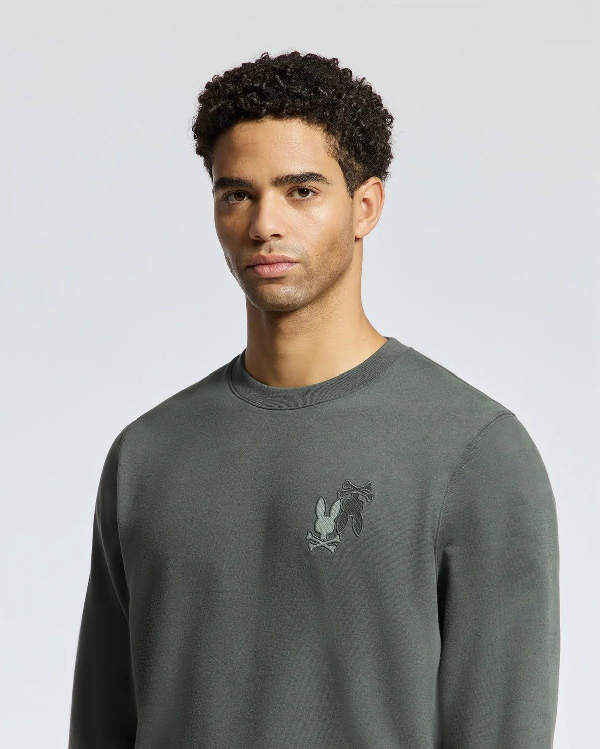 A person with short, curly hair wears the Psycho Bunny Men's Wordmark Peached Terry Crew - B6S278E200 in a plain, dark green, knit cotton style with a small embroidered design on the chest. The background is a neutral gray.