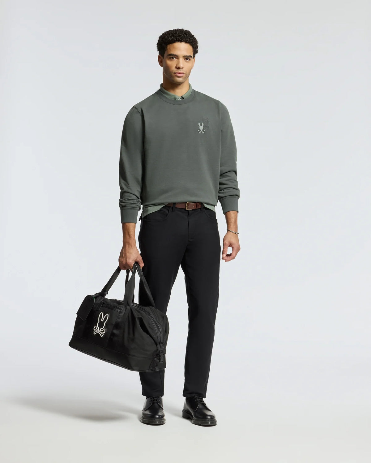 A man wearing a Psycho Bunny MENS WORDMARK PEACHED TERRY CREW in green and black pants is holding a black duffel bag. He has short curly hair and is wearing black shoes, with a plain light color background behind him.