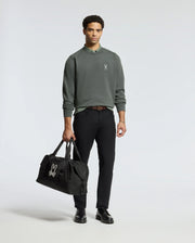 A man wearing a Psycho Bunny MENS WORDMARK PEACHED TERRY CREW in green and black pants is holding a black duffel bag. He has short curly hair and is wearing black shoes, with a plain light color background behind him.