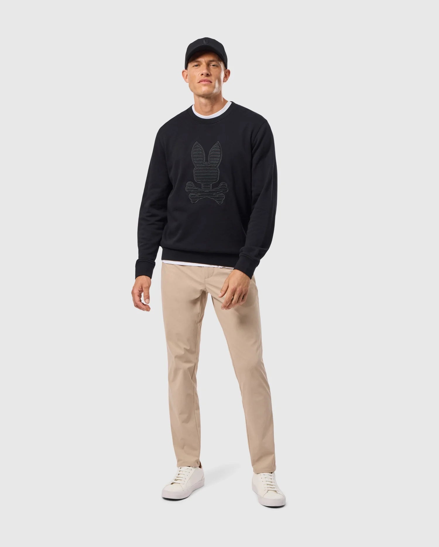 A man stands wearing a black Psycho Bunny MENS KINGSTON SWEATSHIRT - B6S480D200, beige pants, and white sneakers. The lush cotton jersey fabric of the sweatshirt adds comfort and style. His outfit is completed with a black cap against a solid light gray background.
