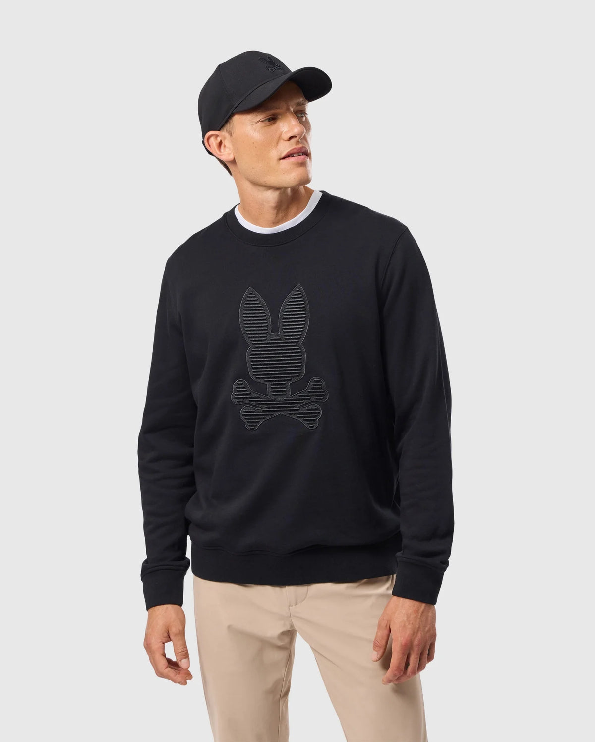 A man sports a black Psycho Bunny MENS KINGSTON SWEATSHIRT - B6S480D200, featuring an eye-catching striped bunny and crossbones design in 3D embroidery. His black baseball cap adds to his outfit, while beige pants complete the look. He gazes to the side against a plain, light background.