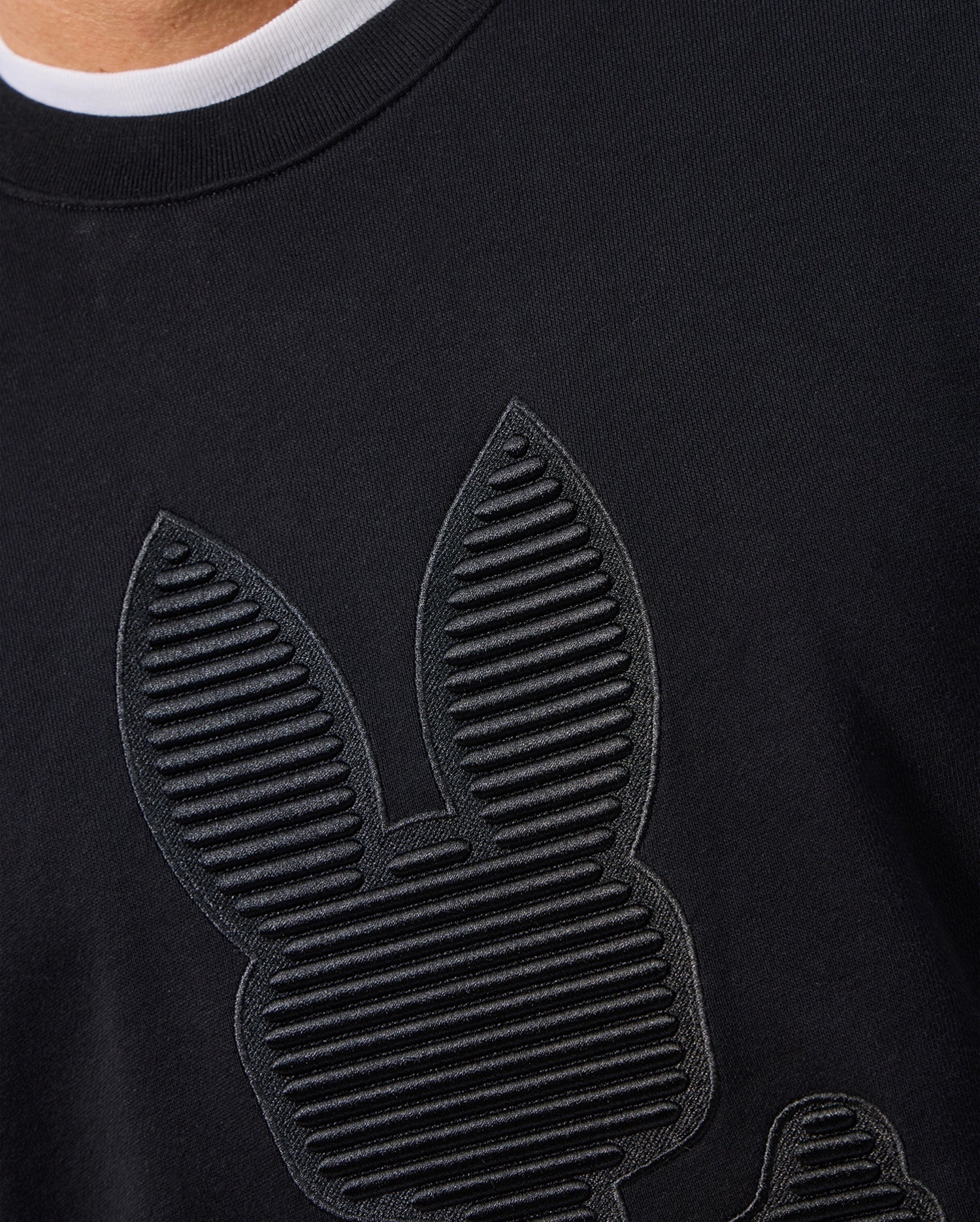 Close-up of a black cotton jersey shirt featuring embossed, vertical line patterns that create bunny ears and a face. The shirt's texture is accentuated by the lighting, with a glimpse of a white neckline at the top, reminiscent of the style seen in the MENS KINGSTON SWEATSHIRT - B6S480D200 by Psycho Bunny.