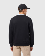 A man wearing the Psycho Bunny MENS KINGSTON SWEATSHIRT - B6S480D200, which features a bold logo with Kingston 3D-embroidery, pairs it with a black cap against a plain gray background. His beige pants highlight the rich cotton jersey material of his striking outfit as he turns away.