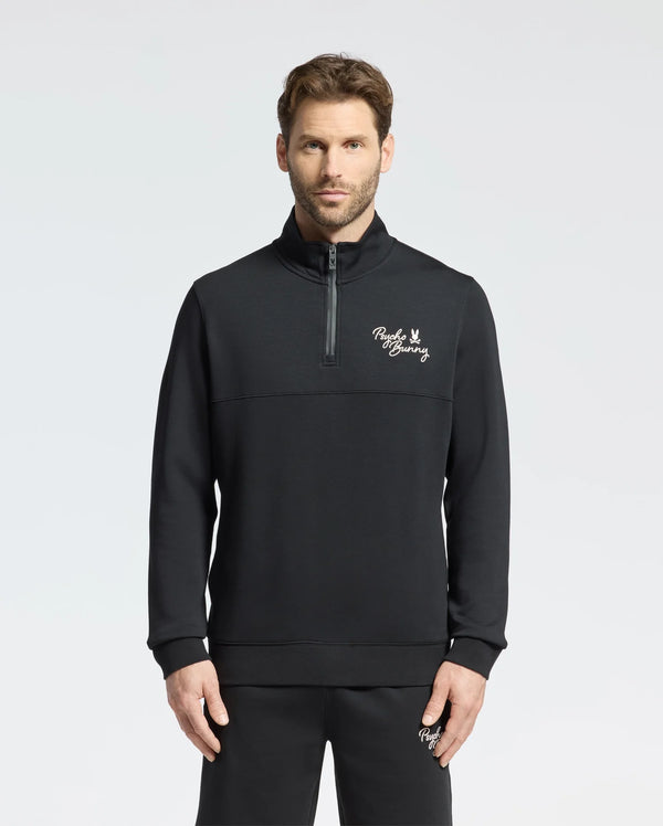A man wearing the Psycho Bunny MENS DESERT 1/4 MOCK ZIP - B6S488F200 stands against a plain background. The sleek black pullover, featuring an embroidered wordmark logo on the chest, is crafted from 100% cotton for a comfortable and stylish casual outfit.