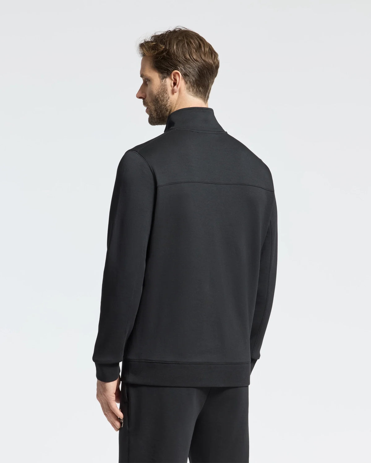 A man with short brown hair stands looking away, donning the Psycho Bunny MENS DESERT 1/4 MOCK ZIP - B6S488F200. This sleek black ensemble features an embroidered wordmark logo and matching pants, with a modern quarter-zip design set against a plain light gray background.