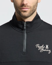 A close-up of a person wearing the MENS DESERT 1/4 MOCK ZIP - B6S488F200 by Psycho Bunny. It’s a black quarter-zip sweatshirt with white embroidered logo on the upper left side. The zipper is partially open, highlighting its sleek design, and the face is partially visible.