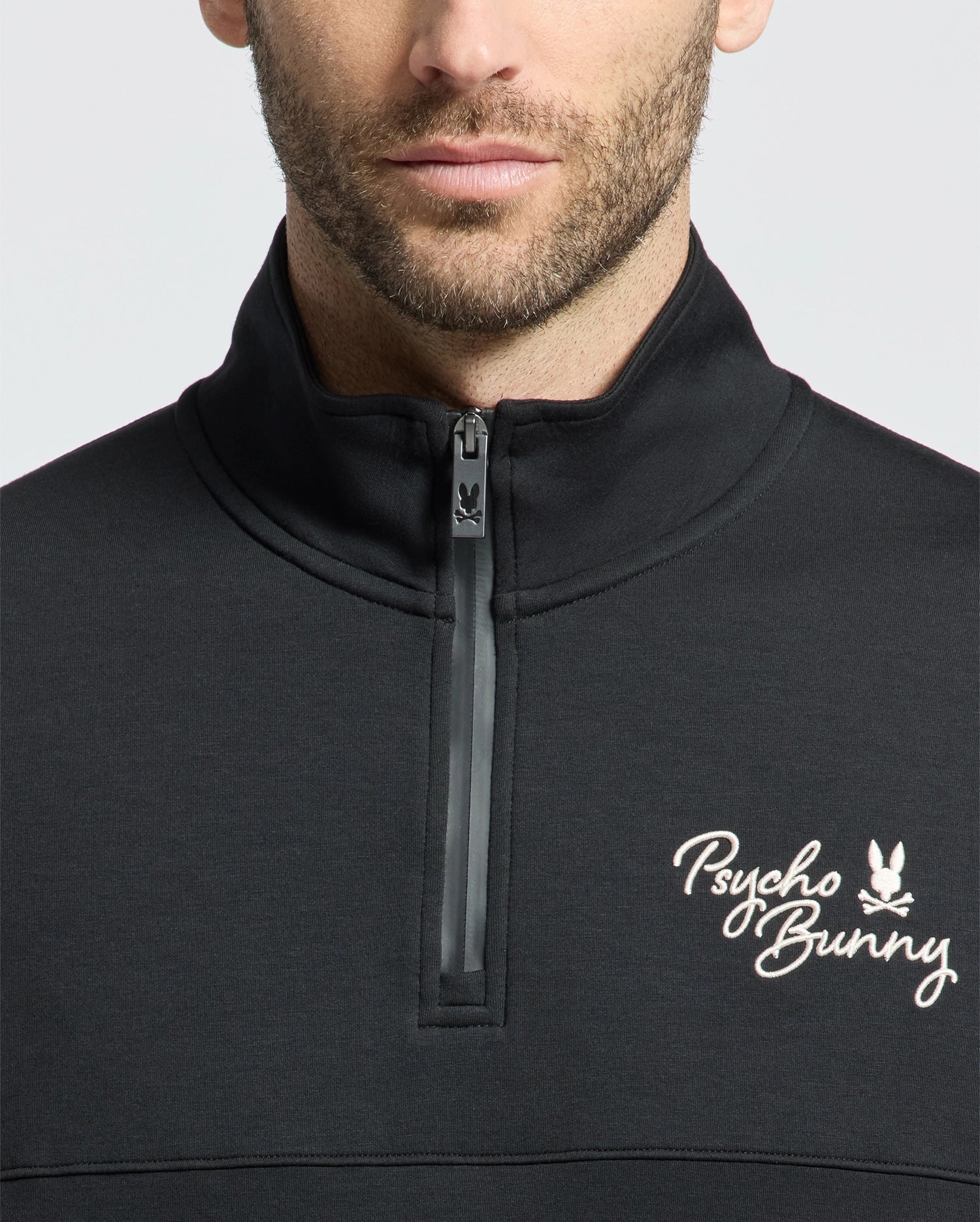A close-up of a person wearing the MENS DESERT 1/4 MOCK ZIP - B6S488F200 by Psycho Bunny. It’s a black quarter-zip sweatshirt with white embroidered logo on the upper left side. The zipper is partially open, highlighting its sleek design, and the face is partially visible.