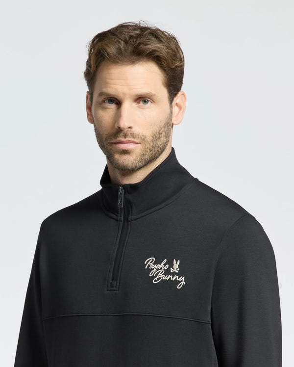 A person with short brown hair and facial hair wears the Psycho Bunny MENS DESERT 1/4 MOCK ZIP, B6S488F200. The black top features a white "Psycho Bunny" embroidered logo on the chest, and a light gray background highlights the stylish 100% cotton design.