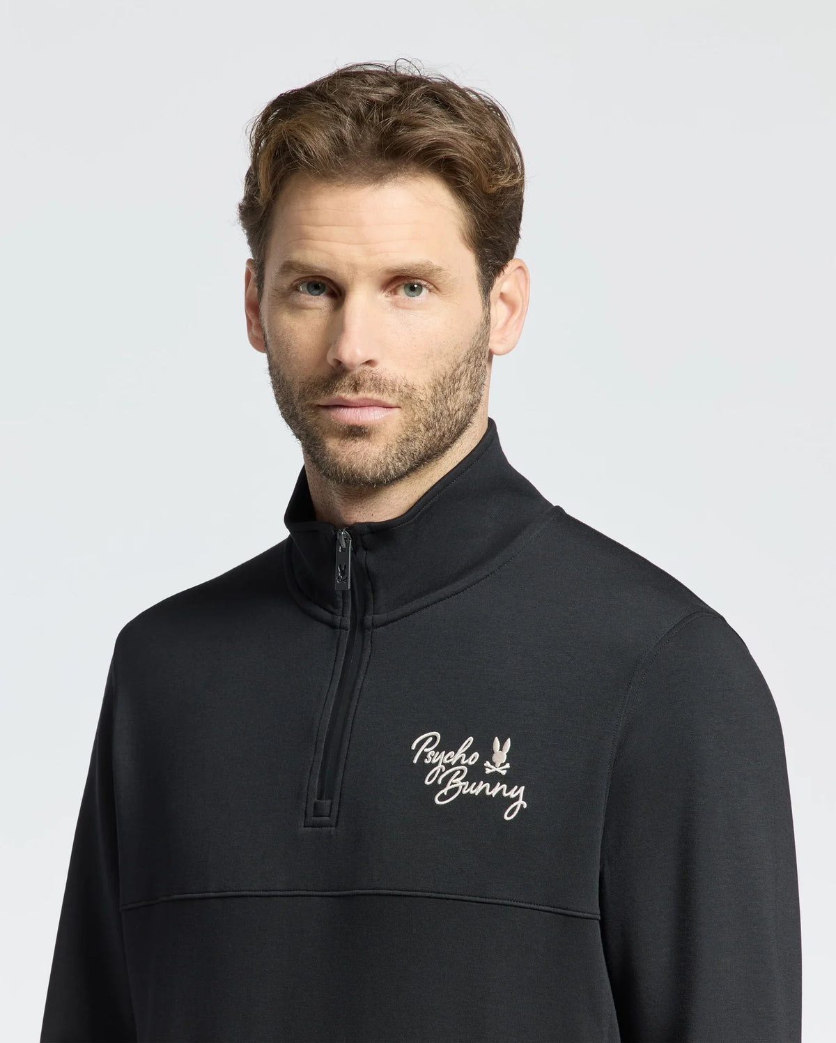 A person with short brown hair and facial hair wears the Psycho Bunny MENS DESERT 1/4 MOCK ZIP, B6S488F200. The black top features a white "Psycho Bunny" embroidered logo on the chest, and a light gray background highlights the stylish 100% cotton design.