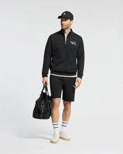 A man wears a Psycho Bunny black sweatshirt and shorts set with an embroidered logo, paired with a matching cap, white socks, and cream sneakers. He holds a large black duffel bag against a plain white background.