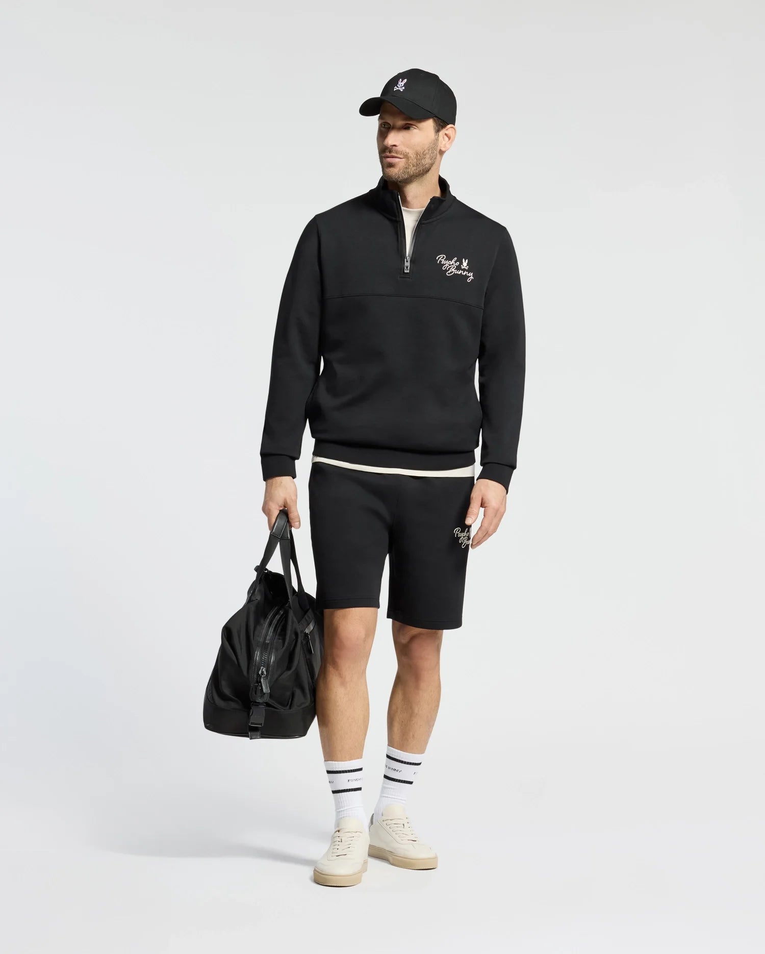 A man wears a Psycho Bunny black sweatshirt and shorts set with an embroidered logo, paired with a matching cap, white socks, and cream sneakers. He holds a large black duffel bag against a plain white background.
