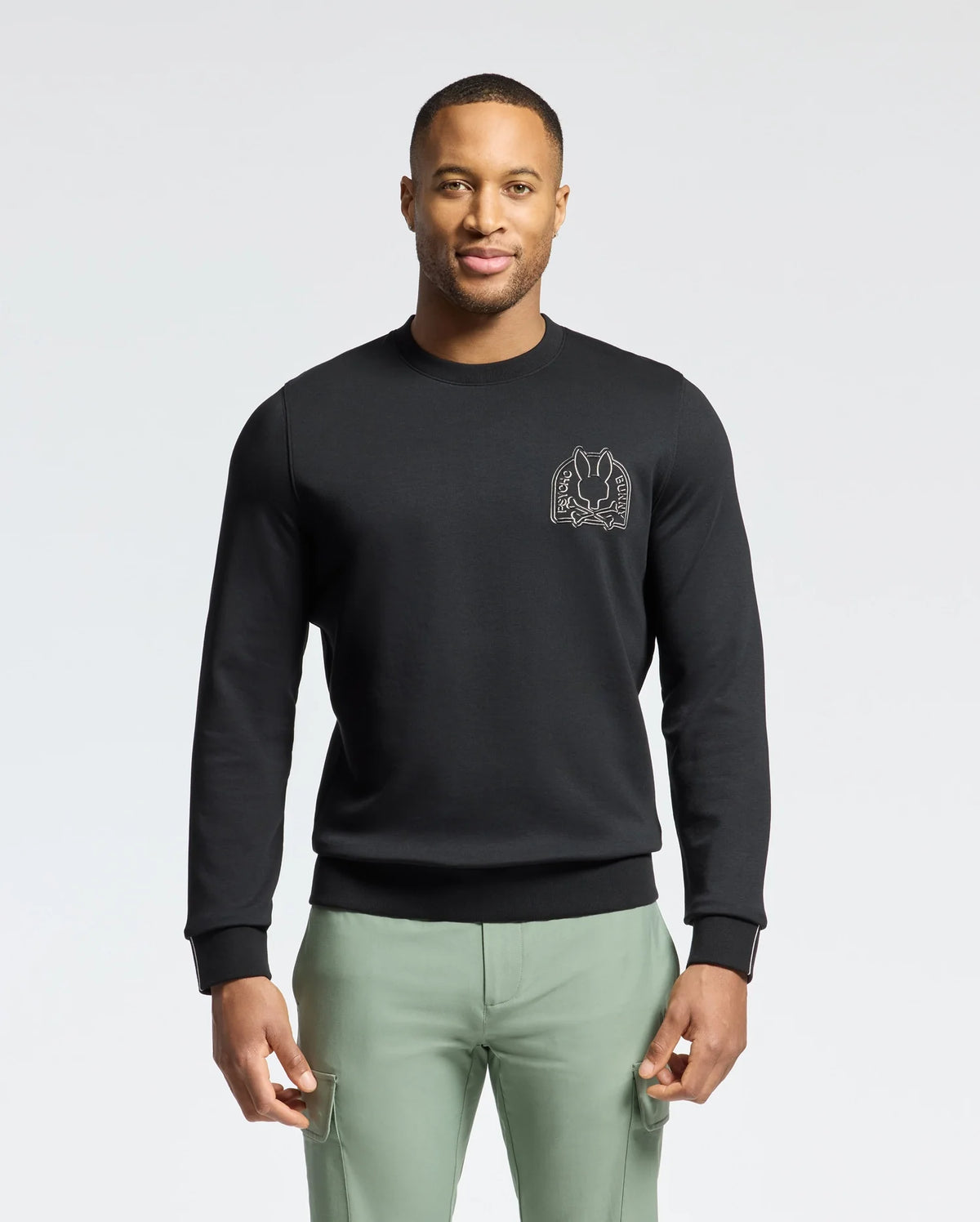 A man wearing a black Psycho Bunny MENS MELBOURNE CLASSIC CREWNECK SWEATSHIRT - B6S561F200 with a small chest graphic stands against a white background. The Modal blend sweatshirt pairs stylishly with his light green pants as he confidently faces the camera.