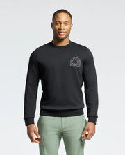 A man wearing a black Psycho Bunny MENS MELBOURNE CLASSIC CREWNECK SWEATSHIRT - B6S561F200 with a small chest graphic stands against a white background. The Modal blend sweatshirt pairs stylishly with his light green pants as he confidently faces the camera.