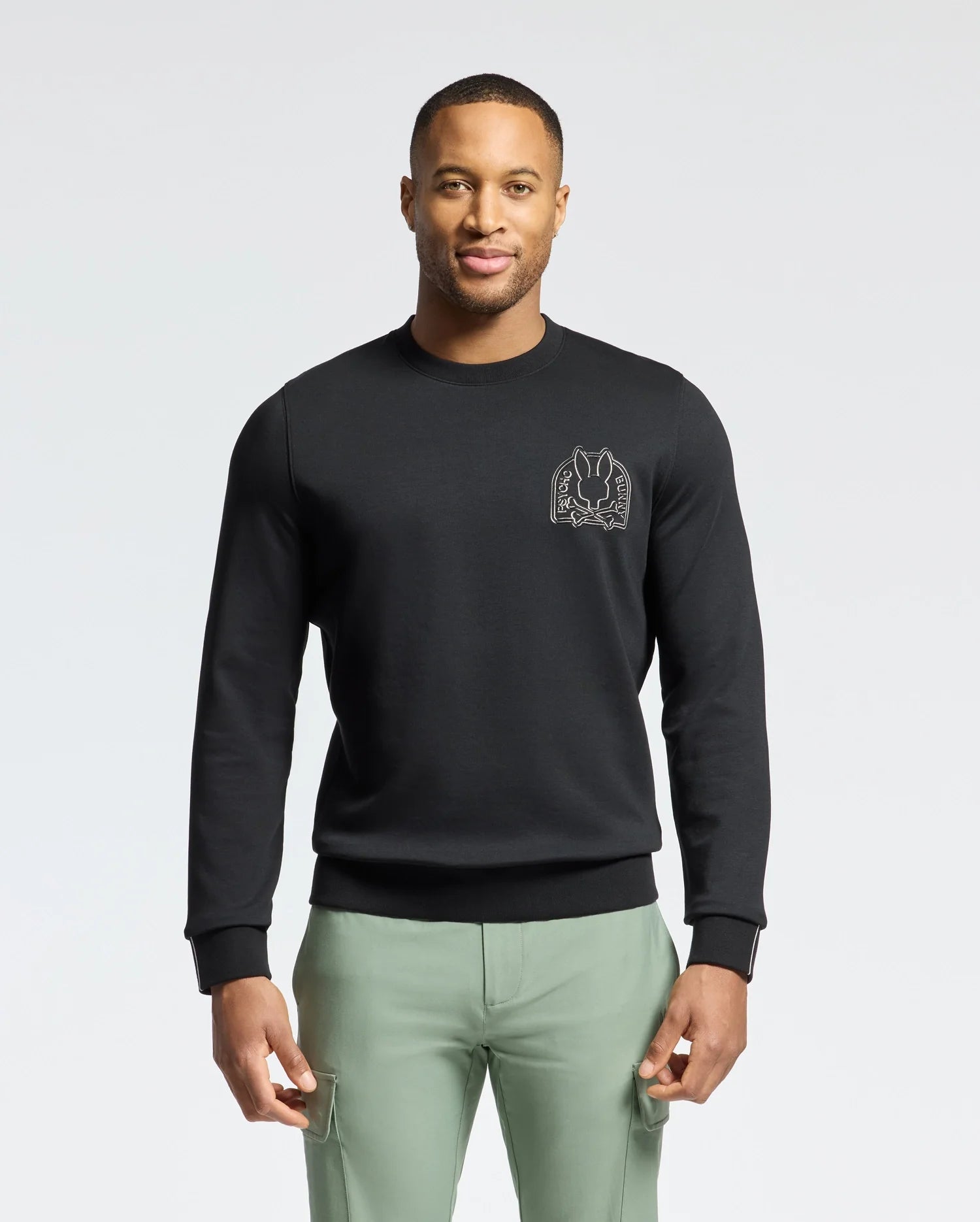 A man wearing a black Psycho Bunny MENS MELBOURNE CLASSIC CREWNECK SWEATSHIRT - B6S561F200 with a small chest graphic stands against a white background. The Modal blend sweatshirt pairs stylishly with his light green pants as he confidently faces the camera.